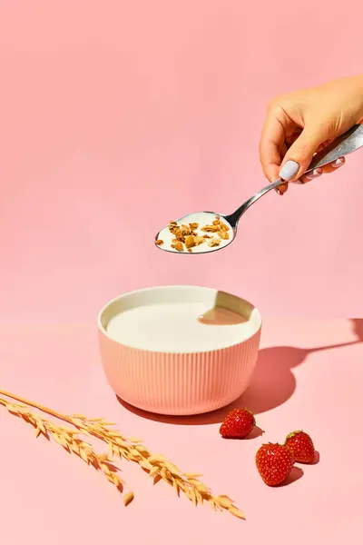 Waman with spoon putting muesli, serial into bowl with warm milk against pink background. Breakfast preparation. Concept of healthy food, nutrition, pop art style, taste. Poster. Copy space for ad