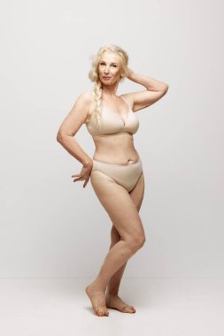 Gri stüdyo arka planında iç çamaşırlarıyla poz veren zarif, güzel, son sınıf öğrencisi bir kadın. Yaş kavramı, doğal güzellik. Vücut ve cilt bakımı, sağlıklı yaşam tarzı.