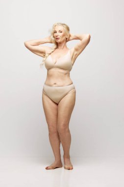 Gri stüdyo arka planında iç çamaşırlarıyla poz veren zarif, güzel, son sınıf öğrencisi bir kadın. Yaş kavramı, doğal güzellik. Vücut ve cilt bakımı, sağlıklı yaşam tarzı.