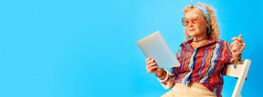 Üst sınıf zarif elbiseli kadın merdivenlerde oturuyor ve mavi stüdyo arka planında tablette haber okuyor. İnsan duyguları, moda, yaşlı insanlar, yaşam tarzı, yaratıcılık kavramı. Ad