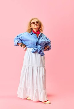 Pembe stüdyo arka planında duran şık etekli ve gömlekli güzel yaşlı kadının tam boy portresi. İnsan duyguları, moda, yaşlı insanlar, yaşam tarzı, yaratıcılık kavramı. Ad
