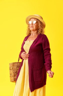 Sarı stüdyo arka planında poz veren güzel, rahat elbiseli, hasır şapkalı ve güneş gözlüklü zarif yaşlı bayan. İnsan duyguları, moda, yaşlı insanlar, yaşam tarzı, yaratıcılık kavramı. Ad