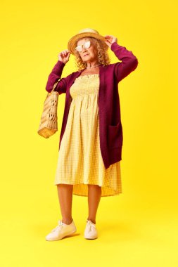Zarif, rahat kıyafetler içinde, sarı stüdyo arka planında poz veren hasır şapkalı ve güneş gözlüklü güzel bir son sınıf hanımı. İnsan duyguları, moda, yaşlı insanlar, yaşam tarzı, yaratıcılık kavramı. Ad