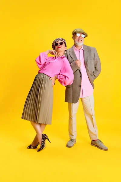 Elegant fashion. Attractive stylish senior woman and man in matching clothes posing against yellow studio background. Concept of beauty and fashion, relationship, modern style, age