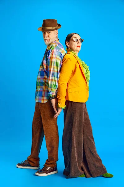 Back to back. Supportive, stylish couple, senior man and woman in fashionable clothes standing against blue studio background. Concept of beauty and fashion, relationship, modern style, age
