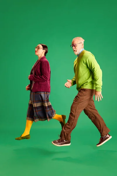 In motion. Senior woman in skirt and jacket walking with man in blazer and pants against green studio background. Concept of beauty and fashion, relationship, modern style, age