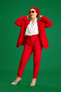 Tam boy. 60 'larında şık, güzel, olgun bir kadın, şık kırmızı bir takım elbise, bere ve güneş gözlüğü takıyor, yeşil stüdyo arka planında duruyor. Güzellik ve moda kavramı, modern tarz, yaş