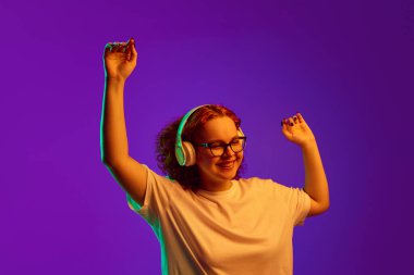 Mutlu, neşeli, gülümseyen genç kız kulaklıkla müzik dinliyor ve neon ışıkta mor arka planda dans ediyor. İnsan duyguları, yaşam tarzı, gençlik, yüz ifadesi kavramı. Ad