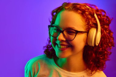 Kıvırcık saçlı, olumlu gülümseyen genç kız kulaklıkla müzik dinliyor. Mor arka planda neon ışıkta. İnsan duyguları, yaşam tarzı, gençlik, yüz ifadesi kavramı. Ad