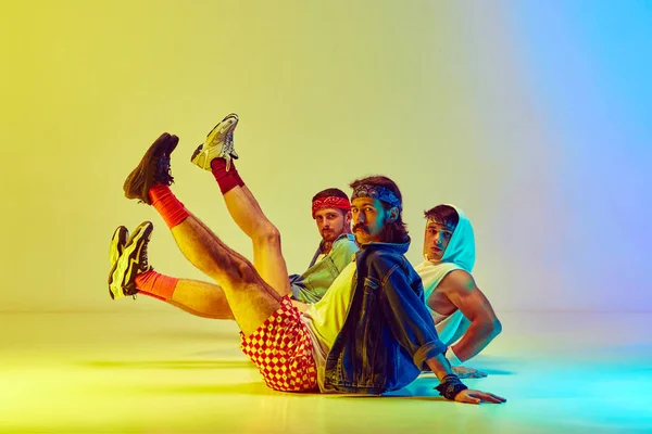 Thee young men, friends in vintage sportswear training, doing aerobics against gradient yellow blue background in neon light. Concept of sportive and active lifestyle, humor, retro style. Ad