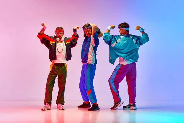 Strength. Three young me, friends wearing colorful old style sportswear, showing muscles against gradient pink blue background in neon light. Concept of sportive and active lifestyle, humor, retro. Ad