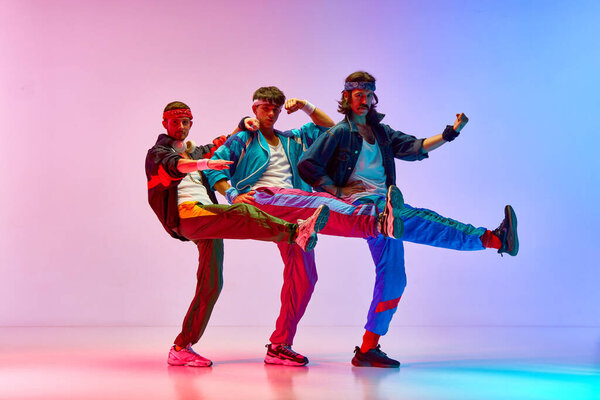 Three men in vintage, funny, colorful sportswear training, stretching against gradient pink blue background in neon light. Concept of sportive and active lifestyle, humor, retro style. Ad