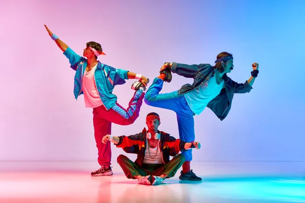 Funny, athletic young men in vintage sportswear training, doing stretching exercises over gradient pink blue background in neon light. Concept of sportive and active lifestyle, humor, retro style. Ad
