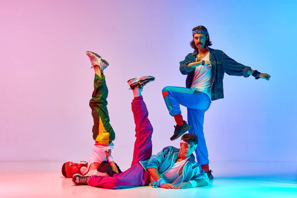 Funny young men in stylish colorful vintage sportswear doing aerobics exercises against gradient pink blue background in neon light. Concept of sportive and active lifestyle, humor, retro style. Ad