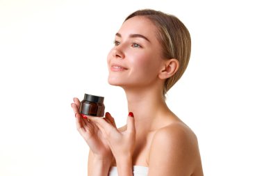 Güzel, gülümseyen genç bir kız beyaz stüdyo arka planında izole edilmiş nemlendirici kremle cildine bakıyor. Doğal güzellik kavramı, cilt bakımı, kozmetik ve kozmetik, spa