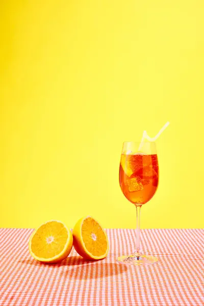 Glass of Aperol spritz cocktail with oranges standing on checkered tablecloth over yellow background. Summer vibe. Concept of alcohol drinks, party, holidays, bar, mix. Poster. Copy space for ad