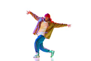 Sportif, şık, spor kıyafetleriyle dans eden sokak dansı yapan genç adam beyaz stüdyo arka planında neon ışıkta izole edilmiş. Çağdaş dans kavramı, sokak tarzı, moda, hobi, gençlik. Ad