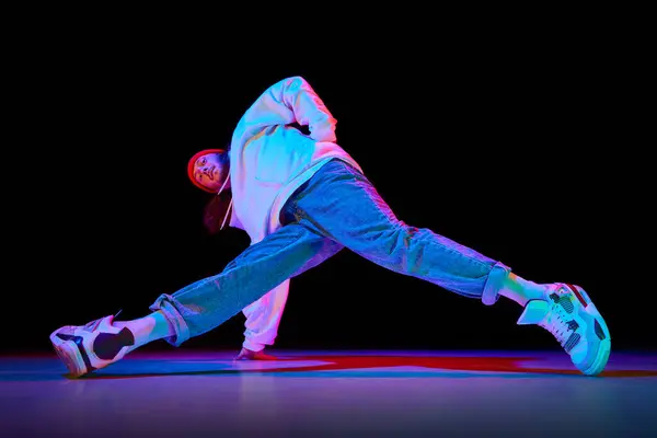 Young man in sportswear in motion, dancing breakdance isolated over black studio background in neon light. Concept of contemporary dance, street style, fashion, hobby, youth. Ad