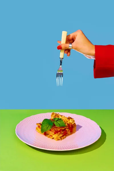 Female hand holding fork over delicious Italian food, lasagna over green and blue background. Bolognese sauce and basil. Concept of Italian food, cuisine, taste, cooking, menu. Pop art. Poster, ad