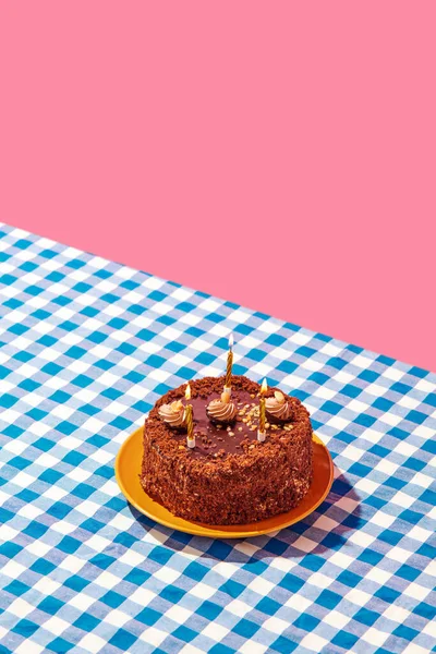 Chocolate delicious cake on plate with candles on blue checkered tablecloth over pink background. Concept of food, desserts, birthday celebration, party. Pop art style. Copy space for ad. Poster