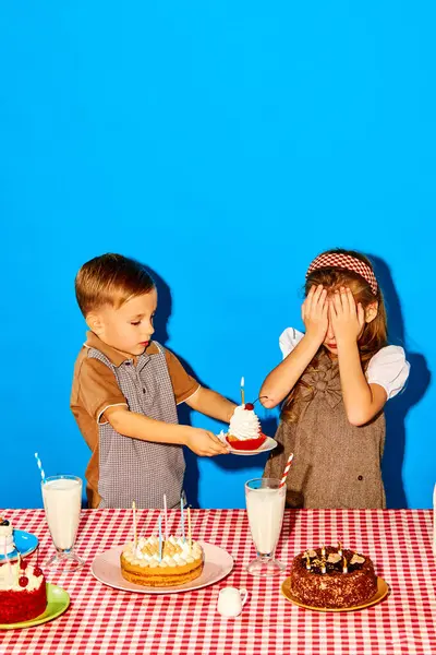 Little boy, brother giving birthday cake with candle to his sister against blue studio background. Siblings birthday. Concept of childhood, birthday celebration, family, fun, food
