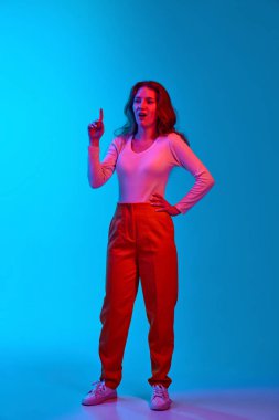 Genç bir kızın gündelik kıyafetlerini giyip parmak kaldırdığı, mavi stüdyonun arka planına karşı parlak fikirler ürettiği uzun bir görüntü. Gençlik kavramı, insan duyguları, yüz ifadesi, yaşam tarzı