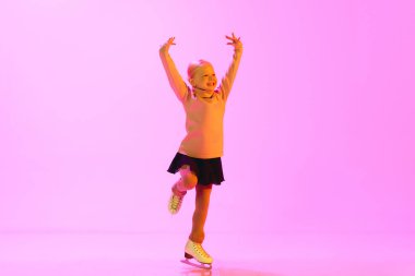 Gülümseyen, mutlu, pozitif küçük çocuk, süveter ve etek giymiş yaşlı kız, pembe arka planda pembe ışıkta dans ediyor. Çocukluk kavramı, artistik patinaj sporu, hobi, okul, eğitim