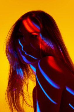 Zarif, düz saçlı, sarı arka planda poz veren güzel bir genç kız. Yüzünde renkli neon ışıklar var. Modern sanat, güzellik, tarz, fütürizm ve siber punk, yaratıcılık kavramı