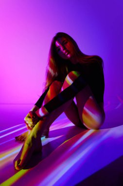 Vücut kıyafetli, mor arka planda oturan, renkli neon ışıkları vücuduna yansıyan şehvetli genç kız. Modern sanat, güzellik, tarz, fütürizm ve siber punk, yaratıcılık kavramı