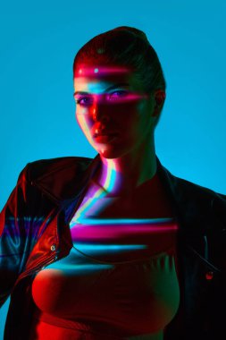 Yüzünde çok renkli neon yansıması olan mavi arka planda kameraya bakan güzel bir genç kadının portresi. Modern sanat, güzellik, tarz, fütürizm ve siber punk, yaratıcılık kavramı