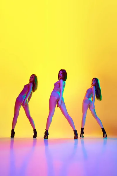 Femininity. Three young woman in underwear and heeled shoes dancing high heel dance over gradient yellow background in neon. Concept of hobby, contemporary dance, art, beauty, creativity, elegance