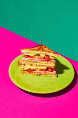 Izgara ekmek, jambon, marul, peynir, domates ve avokadoyla kaplı bir tabak pembe yeşil arka plan. Fast food, tat, yemek, malzeme kavramı. Tamamlayıcı renkler. Poster, reklam
