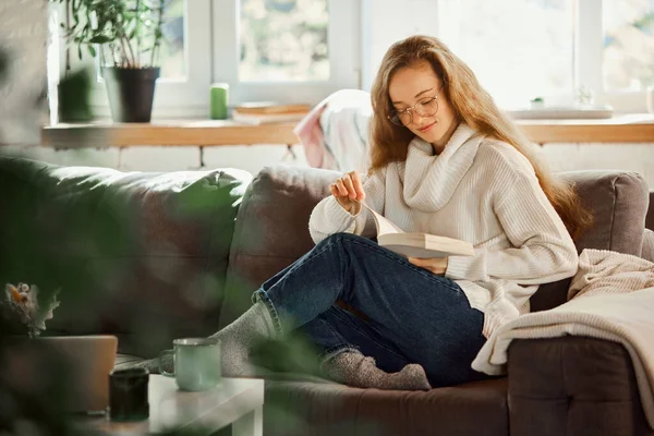 Young beautiful woman sitting on coach and reading book. Full length shot of happy young woman relaxing at home. concept of lifestyle, winter holiday season, autumn weekend, relax and cozy atmosphere.