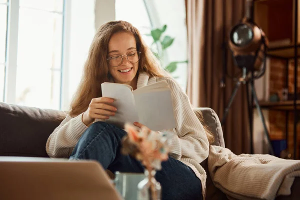Happy cheerful young woman sitting and reading book on coach at home in quiet and cozy atmosphere. Concept of lifestyle, winter holiday season, autumn weekend, relax and cozy atmosphere,