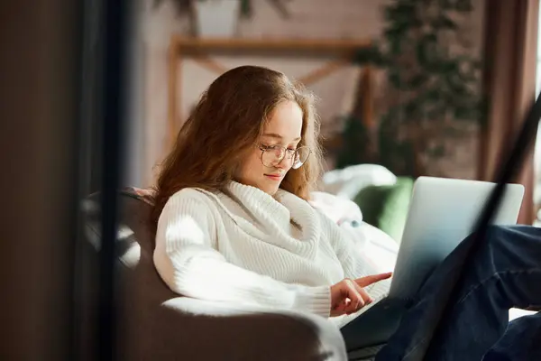 Cute cozy young woman sitting in soft comfortable armchair and watching movies in laptop on Christmas Eve. concept of lifestyle, winter holiday season, autumn weekend, relax and cozy atmosphere.