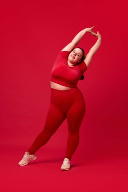 Rahat spor giyim eğitimi almış, kırmızı stüdyo arka planına yaslanmış kilolu güzel bir kadın. Spor anlayışı, vücut pozitifliği, kilo kaybı, vücut ve sağlık hizmetleri, kendini kabul etme