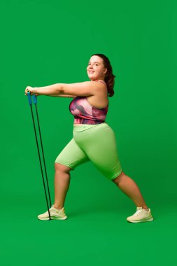 Genç aşırı kilolu, spor giyim eğitiminde şişman bir kadın yeşil stüdyo geçmişine karşı fitness genişletici egzersizler yapıyor. Spor anlayışı, vücut pozitifliği, kilo kaybı, vücut ve sağlık hizmetleri