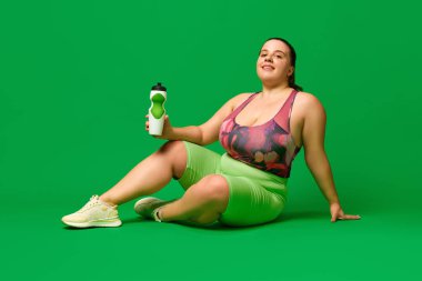 Sıvı kaybı. Büyük beden manken, spor giyimli şişman bir kadın yeşil stüdyo arka planında su şişesiyle yerde oturuyor. Spor anlayışı, vücut pozitifliği, kilo kaybı, vücut, sağlık hizmetleri