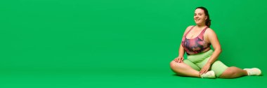 Büyük beden manken, spor giyimli şişman bir kadın yeşil stüdyo arka planında yerde oturuyor. Spor anlayışı, vücut pozitifliği, kilo kaybı, vücut ve sağlık hizmetleri. Pankart