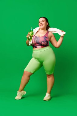 Sıvı kaybı. Büyük beden model, spor giyimli şişman bir kadın su şişesi ve havluyla yeşil stüdyo arka planında duruyor. Spor anlayışı, vücut pozitifliği, kilo kaybı, vücut, sağlık hizmetleri.