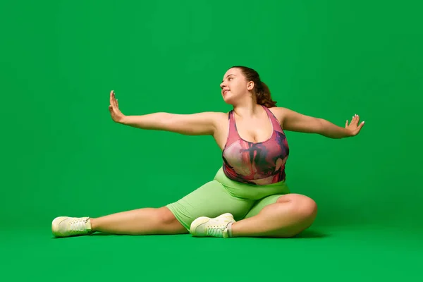 Body stretch. Fat, overweight, chubby woman training, doing exercises against green studio background. Concept of sport, body-positivity, weight loss, body and health care