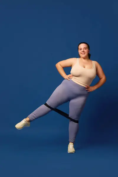 Leg swings. Young overweigh woman training with fitness elastic band against blue studio background. Concept of sport, body-positivity, weight loss, body and health care. Copy space for ad