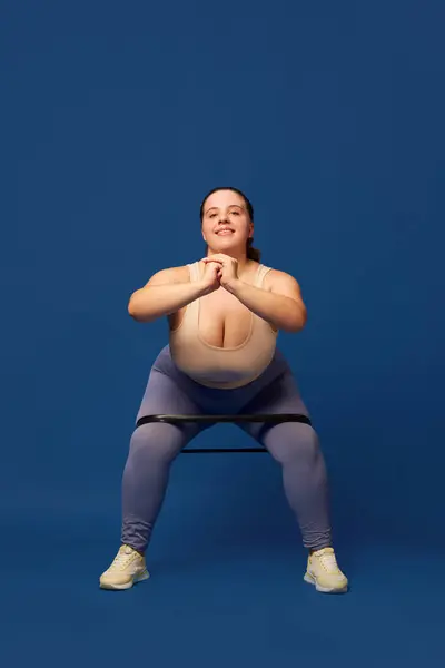 Young overweigh woman training with fitness elastic band, doing squats against blue studio background. Concept of sport, body-positivity, weight loss, body and health care. Copy space for ad