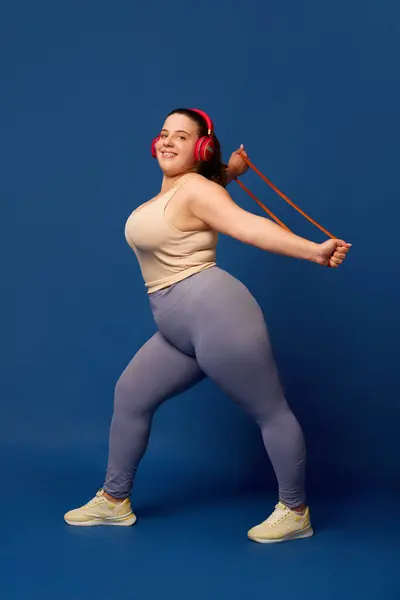 Smiling young woman with fat body training with elastic bands, fitness expanders against blue studio background. Concept of sport, body-positivity, weight loss, body and health care. Copy space for ad