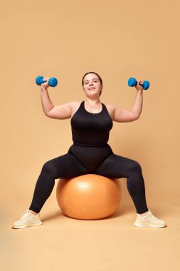 Spor giyimli, spor kıyafetli, spor topu ve halterli genç bir kadın bej renkli stüdyo arka planına karşı. Spor anlayışı, vücut pozitifliği, kilo kaybı, vücut ve sağlık hizmetleri. İlan için alanı kopyala