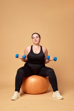 Spor giyimli, spor kıyafetli, spor topu ve halterli genç bir kadın bej renkli stüdyo arka planına karşı. Spor anlayışı, vücut pozitifliği, kilo kaybı, vücut ve sağlık hizmetleri. İlan için alanı kopyala