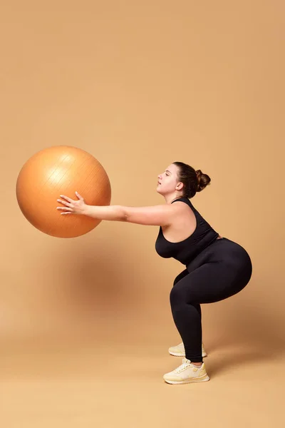 Young overweigh woman in black sportswear training with fitness ball, doing squats against beige studio background. Concept of sport, body-positivity, weight loss, body, health care. Copy space for ad