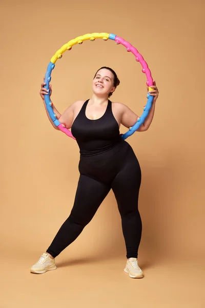 Overweight young woman in black sportswear training with hoop against beige studio background. Concept of sport, body-positivity, weight loss, body and health care. Copy space for ad
