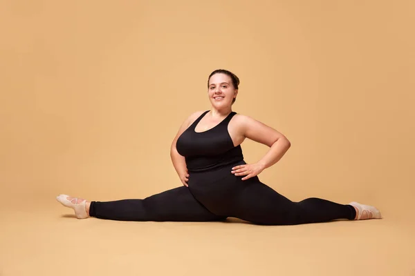 Young overweight woman in black sportswear training, sitting on twine against beige studio background. Concept of sport, body-positivity, weight loss, body and health care. Copy space for ad