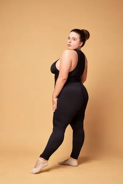 Beautiful young overweight woman in black sportswear standing against beige studio background. Concept of sport, body-positivity, weight loss, body and health care. Copy space for ad